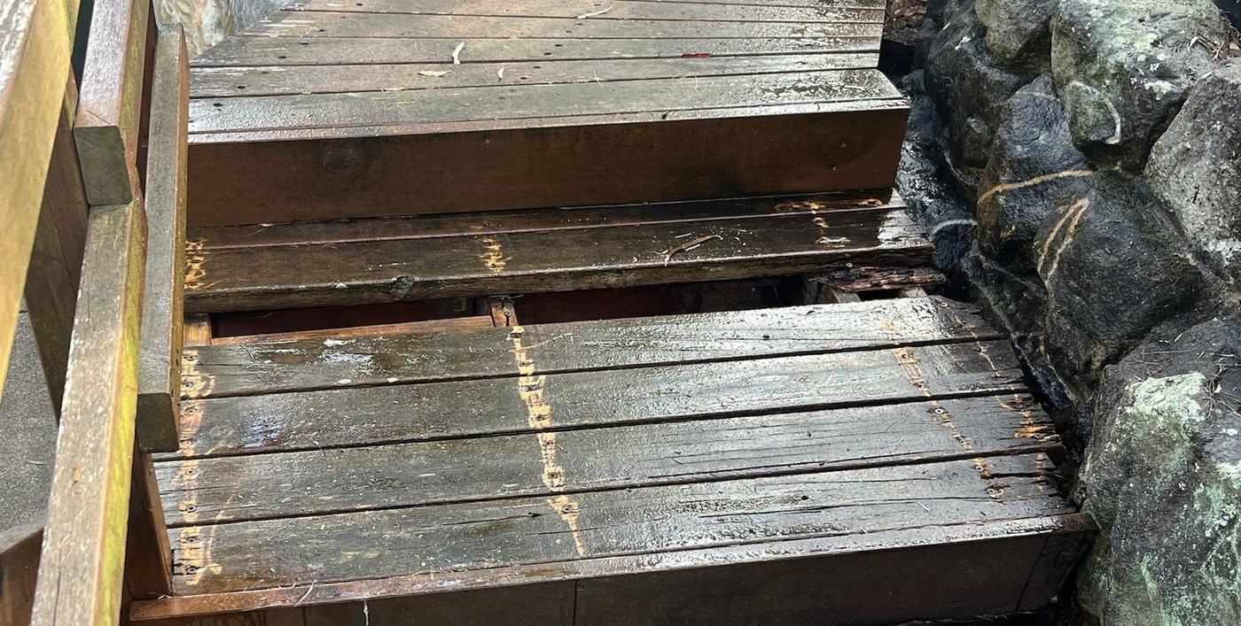 Close-up of a rotten timber stair tread and riser pressed against a sandstone wall, split and breaking away at the edge